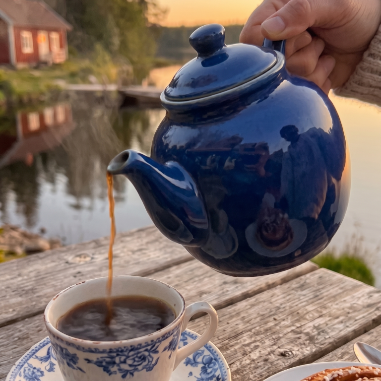 En blå kaffekanna häller upp kaffe i en klassisk porslinskopp på ett träbord utomhus, med en röd svensk stuga vid en sjö i bakgrunden.